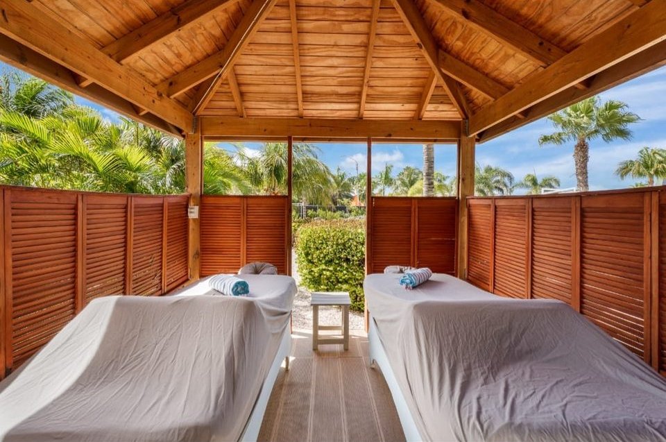 Open-air treatment cabana with a pitched wooden ceiling and twin massage beds, framed by tropical planting and blue sky.