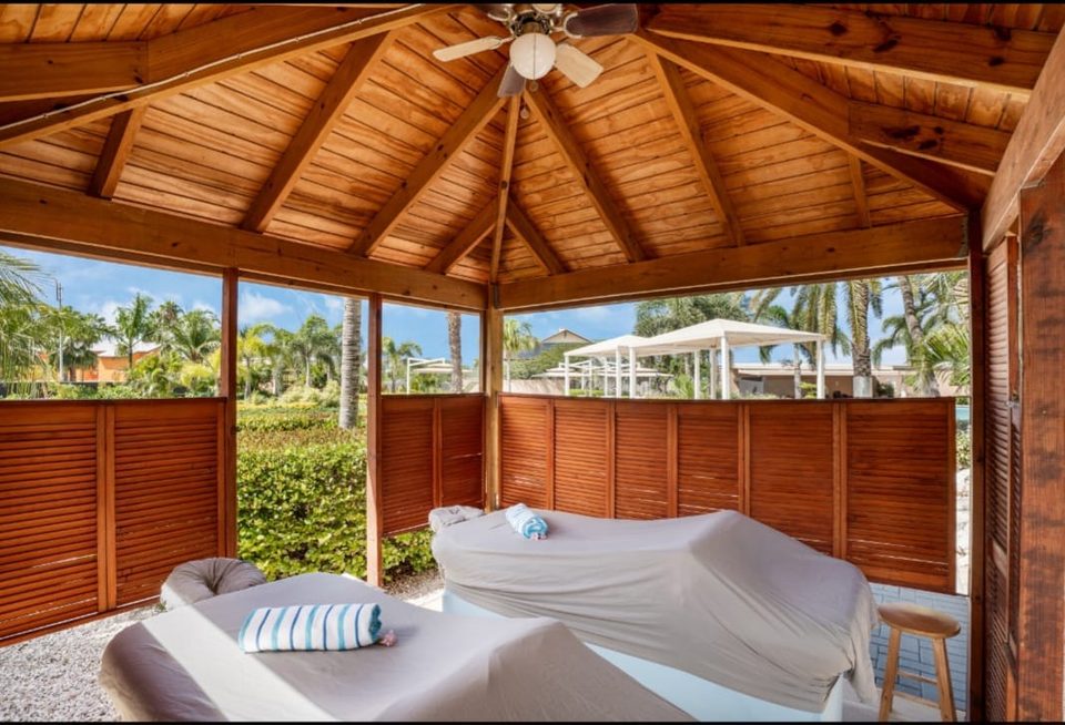 Two massage beds made up with white linens inside an open wooden cabana at Le Vent, looking out through louvered screens to palms and garden.