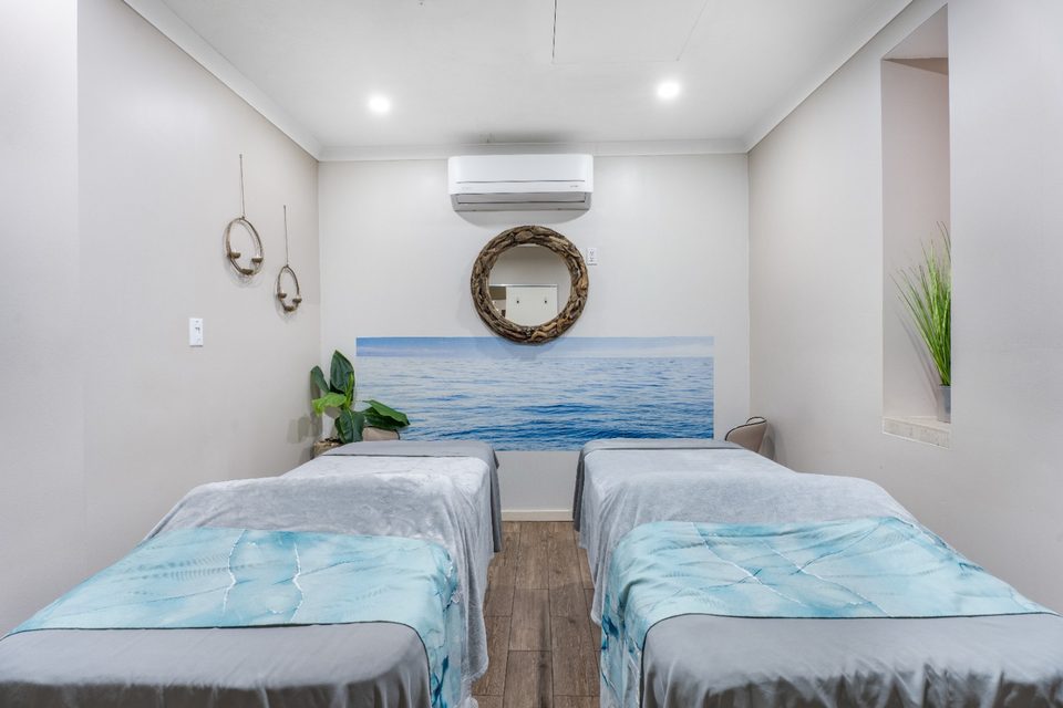 Couples treatment room with two beds dressed in turquoise and white linens beneath a round mirror and a painted wave mural.