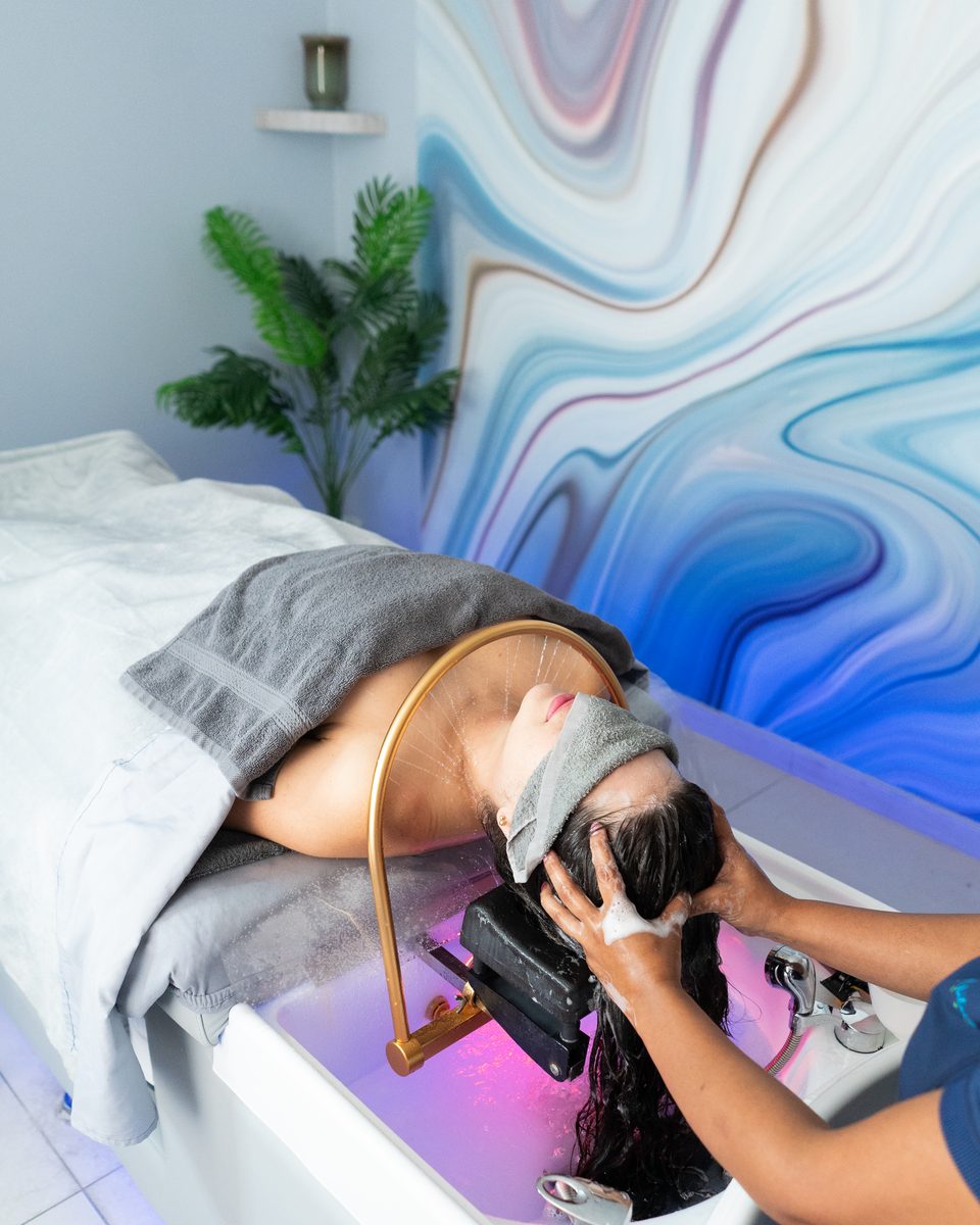 Therapist rinsing a guest's hair during a head spa treatment in a room with a flowing marbled mural.