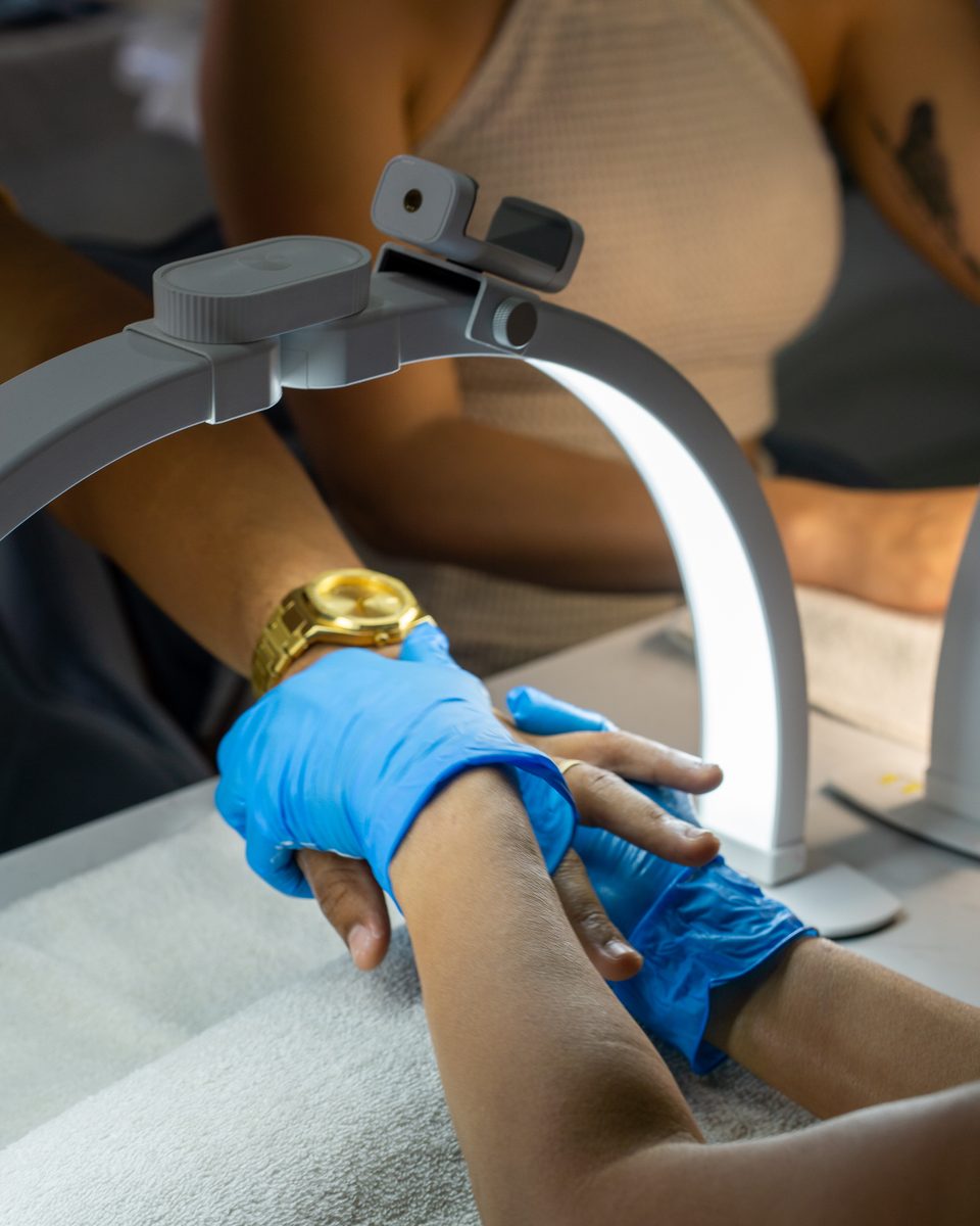 Nail technician in blue gloves preparing a guest's hand under a ring lamp.