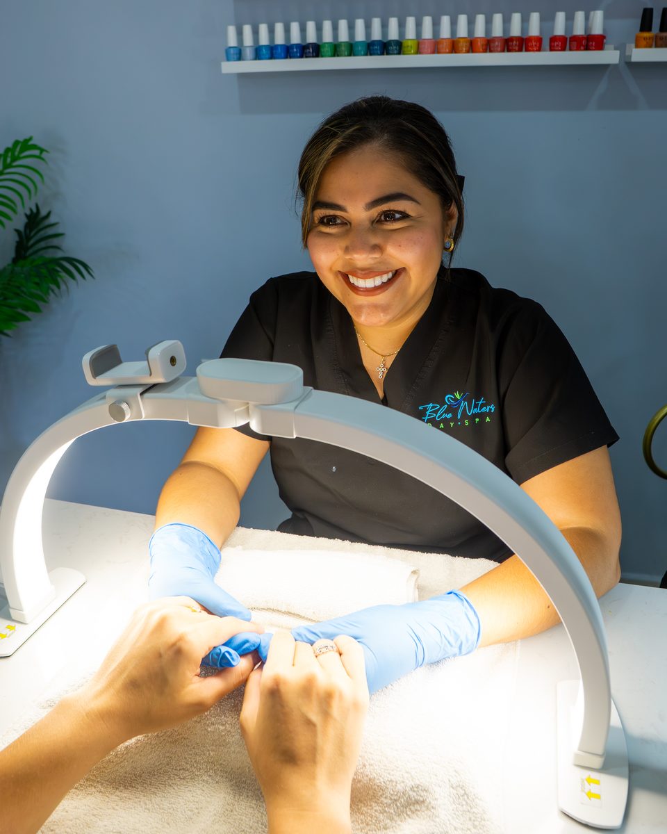 Nail technician at work under a ring lamp, smiling toward the camera.