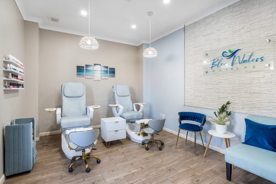 Pedicure studio with white spa chairs, a blue accent sofa and the Blue Waters Day Spa logo on the wall.