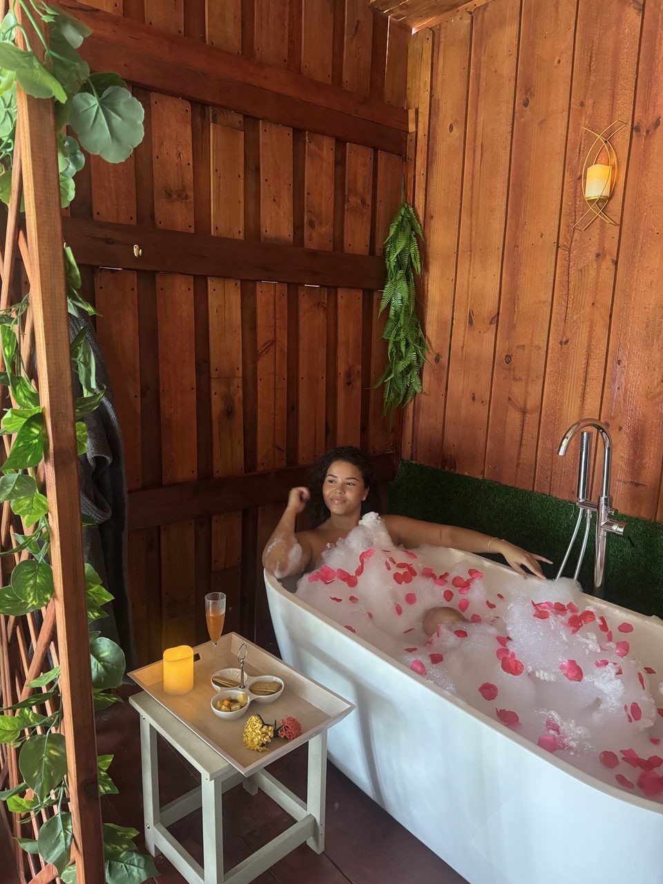 Guest relaxing in a warm aromatic bath with rose petals, a candle and a tray of snacks beside the tub.